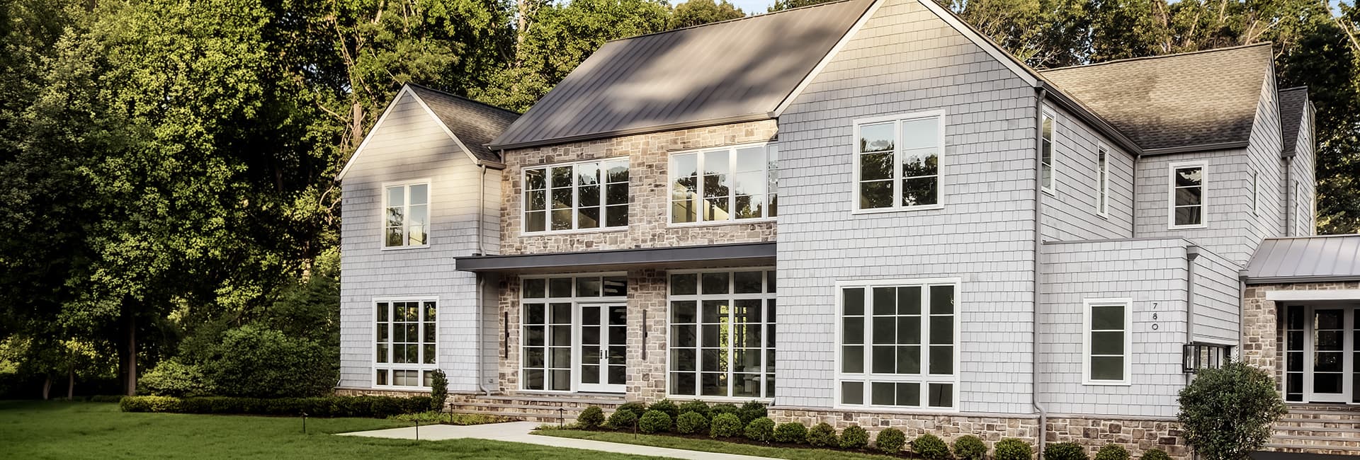 Two-story modern farmhouse exterior with gray shingle siding, stone facade, large windows and landscaped lawn