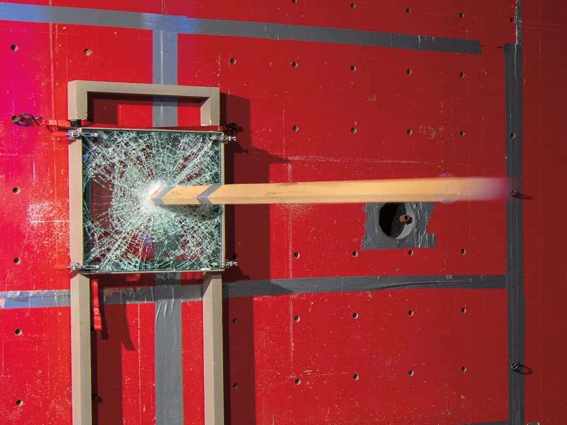 Shattered square safety glass in a frame pierced by a motion-blurred wooden beam during an impact test on a red taped test wall