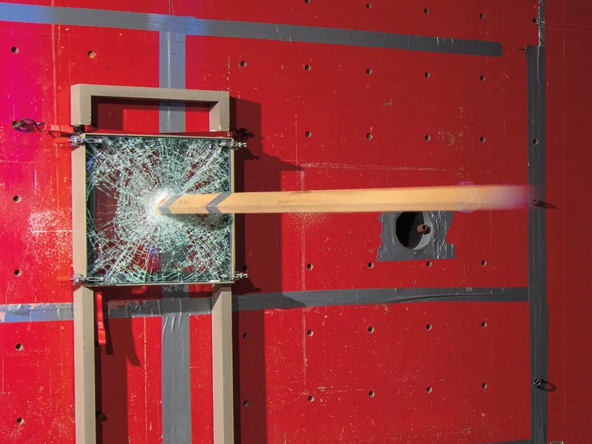 Shattered square safety glass in a frame pierced by a motion-blurred wooden beam during an impact test on a red taped test wall