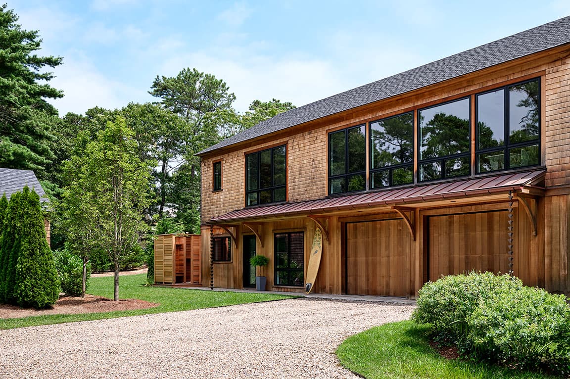 A wooden house on Cape Cod featuring black windows and a gravel driveway, surrounded by greenery.