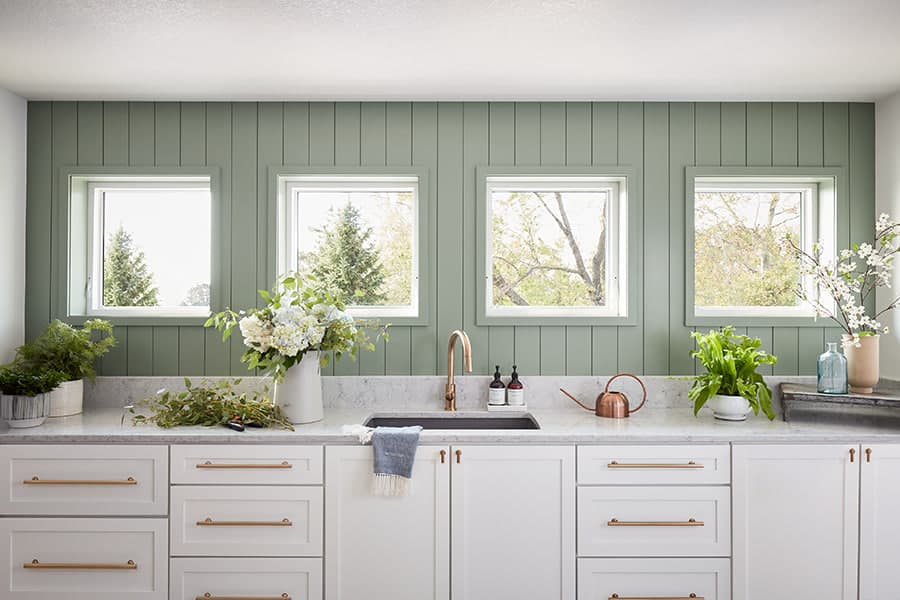 A modern kitchen with a green wall, white cabinets, and multiple picture windows showcasing greenery outside.
