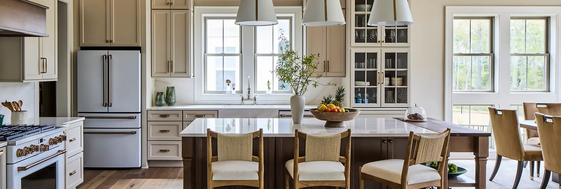 Bright modern kitchen with white island, wooden bar stools, stainless appliances, pendant lights, and large windows