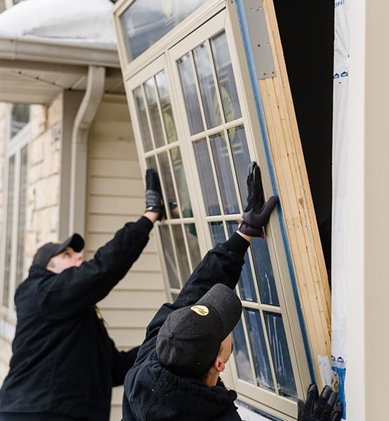 Two installers positioning a large replacement window into an exterior wall during winter