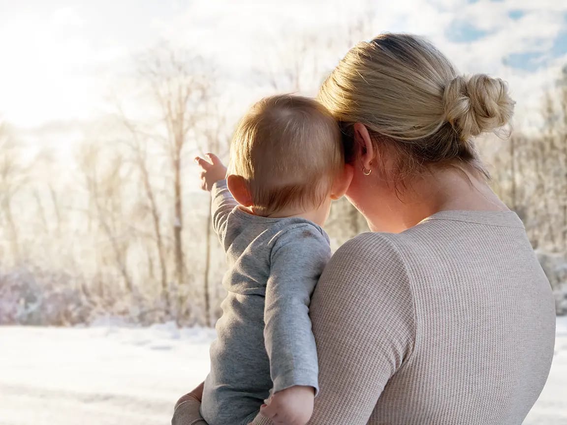 Mother holding baby pointing toward snowy winter trees, back view