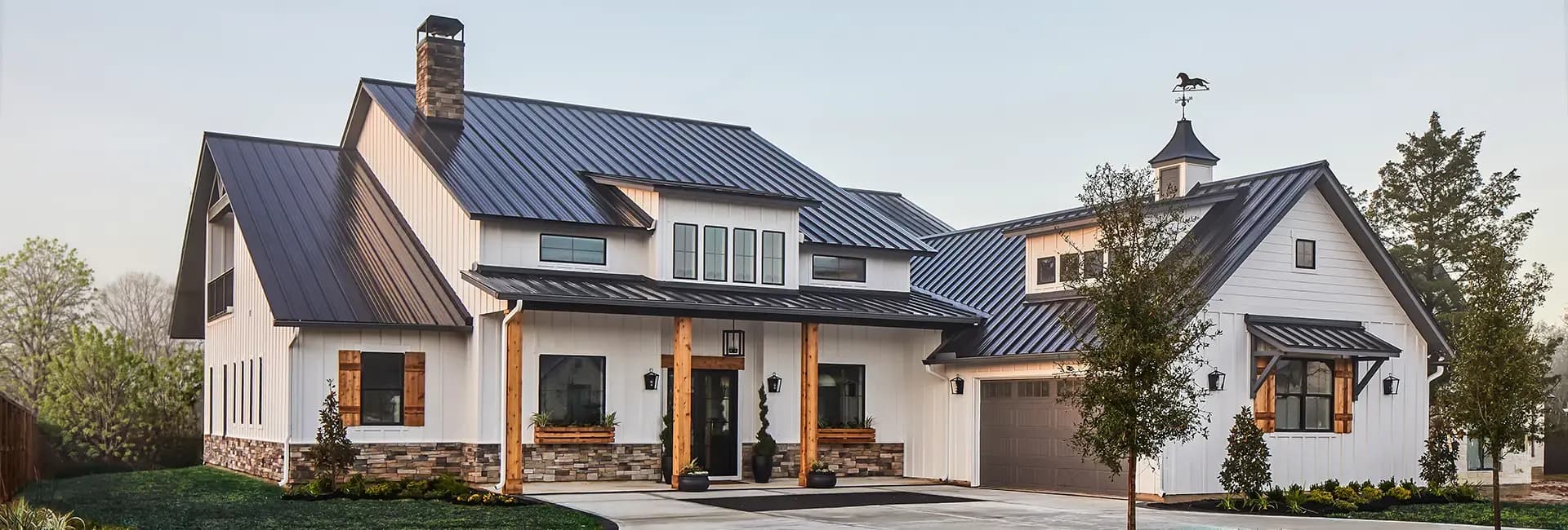Modern white farmhouse with black metal roof, wood accents and stone veneer base, cupola with weathervane, front porch and attached garage