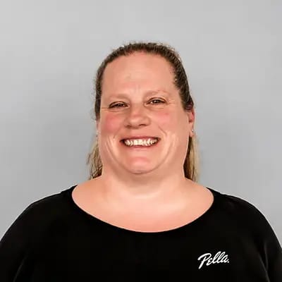 a woman wearing a black Pella shirt is smiling and standing in front of a gray wall