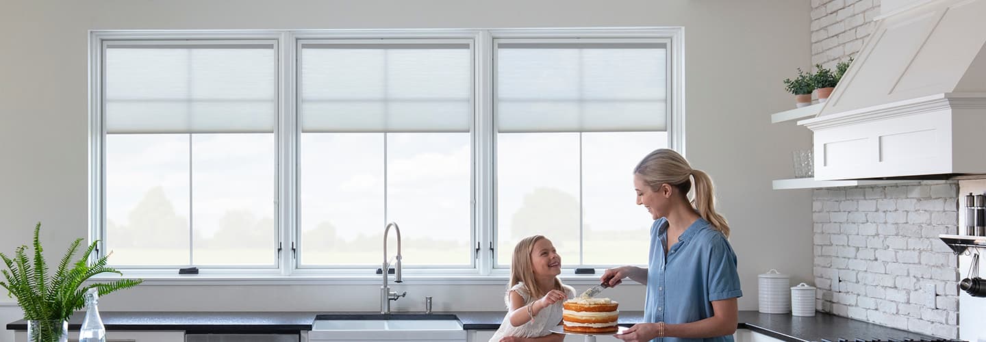 A mother and her daughter are standing in front of three while casement windows decorating a cake.