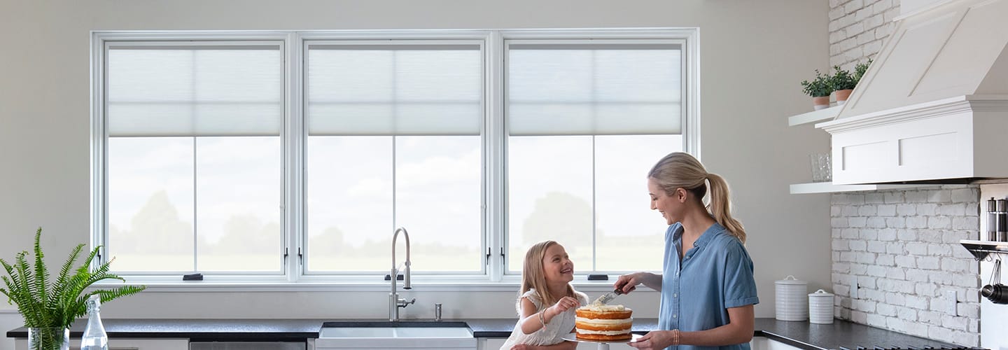 A mother and her daughter are standing in front of three while casement windows decorating a cake.