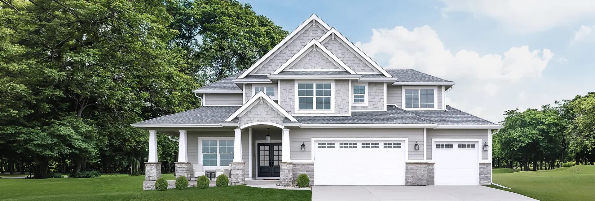 Two-story gray shingle and siding suburban house with a covered front porch, columns, three-car garage, driveway and landscaped lawn with trees
