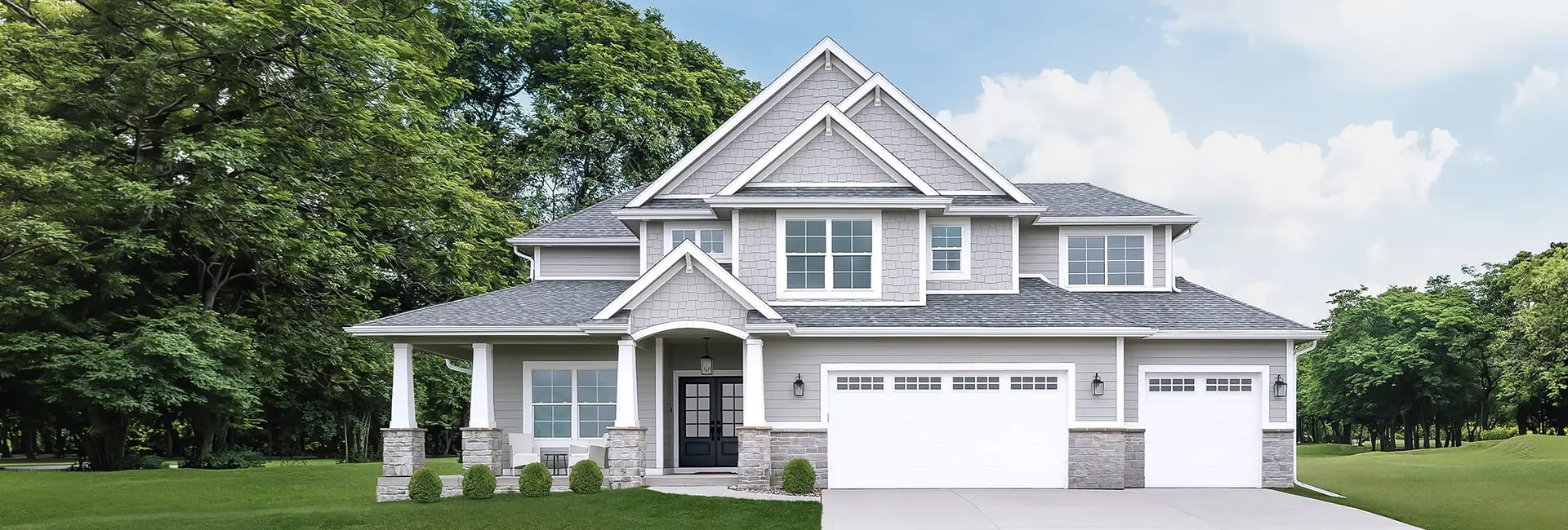 Two-story gray shingle and siding suburban house with a covered front porch, columns, three-car garage, driveway and landscaped lawn with trees