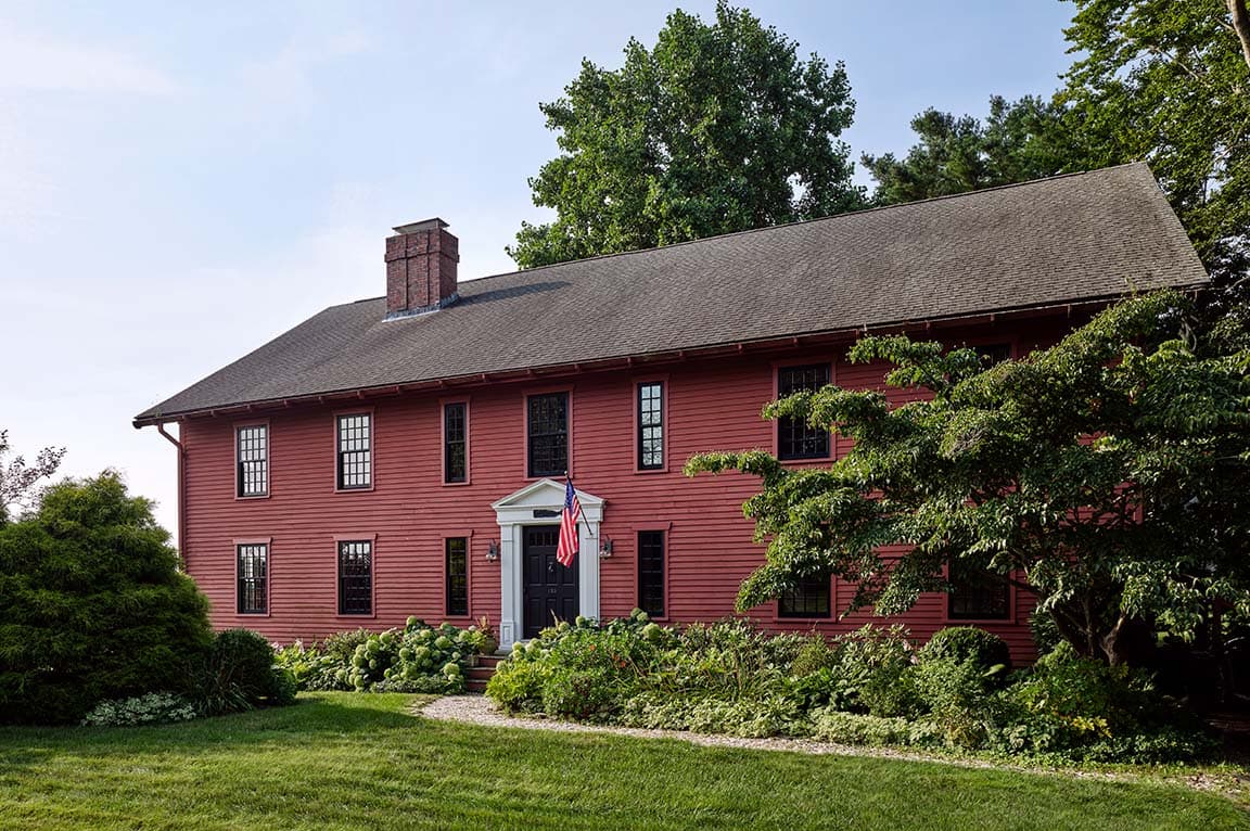 A historic red house with wood windows surrounded by lush greenery and a well-maintained garden.
