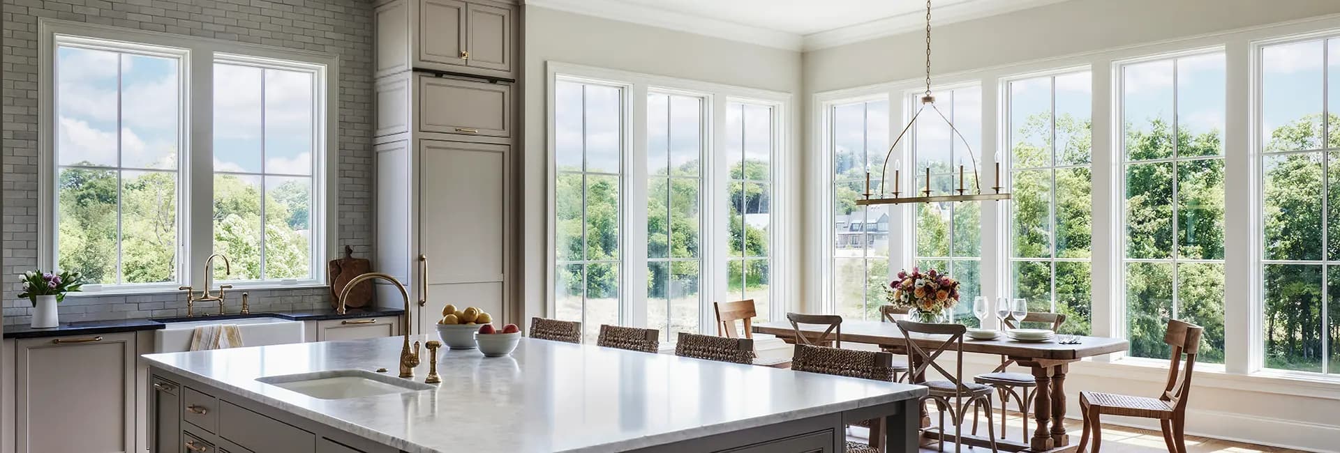 Bright modern farmhouse kitchen with marble island, brass faucet, farmhouse sink and dining table by large windows overlooking green trees.
