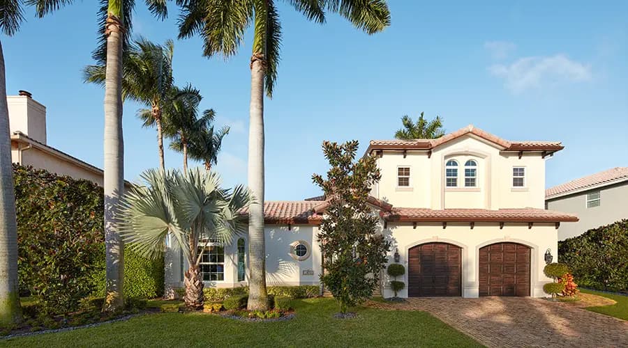 Mediterranean-style two-story home with red tile roof, double wooden garage doors, manicured lawn and palm trees in a tropical front yard under a clear blue sky
