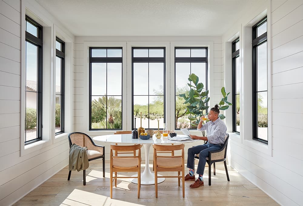A man is sitting at a dining room table surrounded by seven tall black windows.