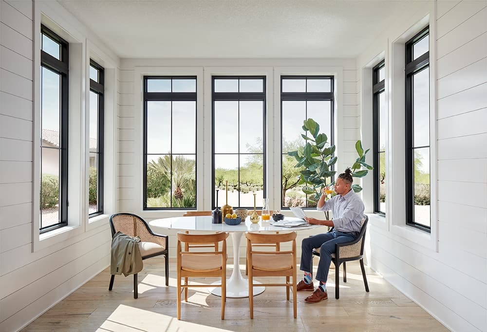 A man is sitting at a dining room table surrounded by seven tall black windows.