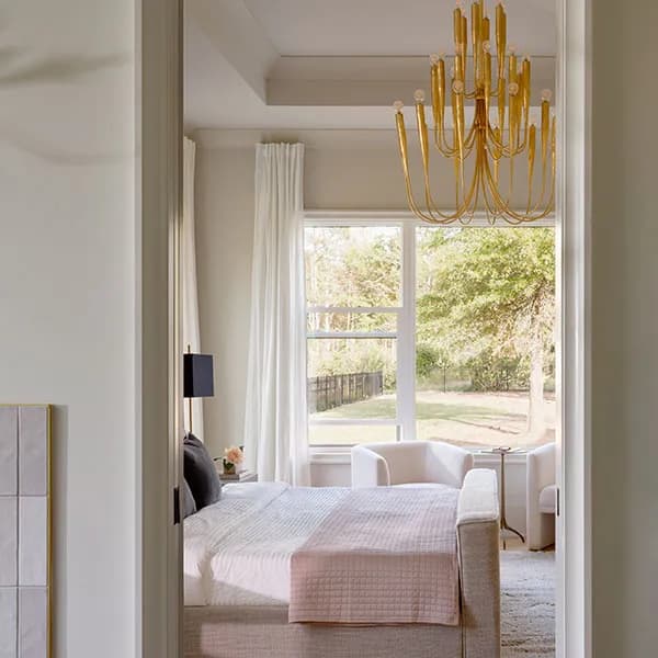 A modern bedroom features combinations of a gold chandelier, large windows, and neutral furnishings.