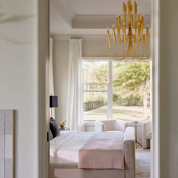 A modern bedroom features combinations of a gold chandelier, large windows, and neutral furnishings.