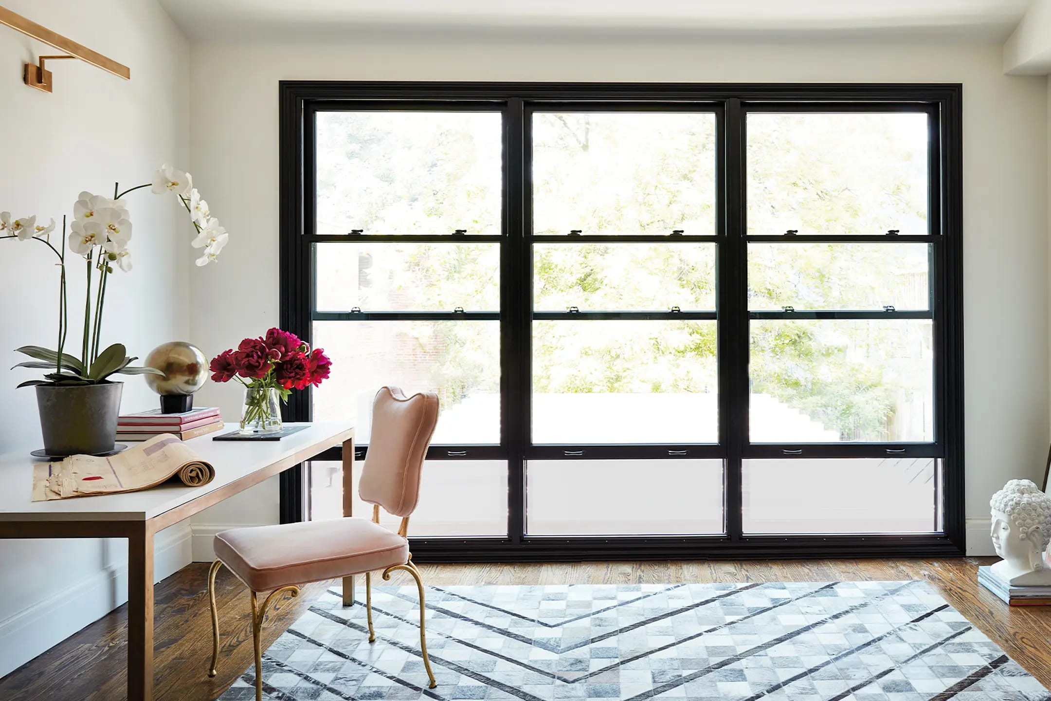 Bright home office with wooden desk, blush-pink velvet chair, orchids and red flowers, large black-framed windows and patterned rug.