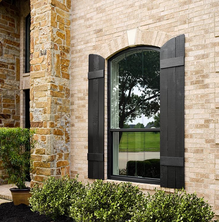 Single black arched double-hung window with black shutters on each side on a brick home.