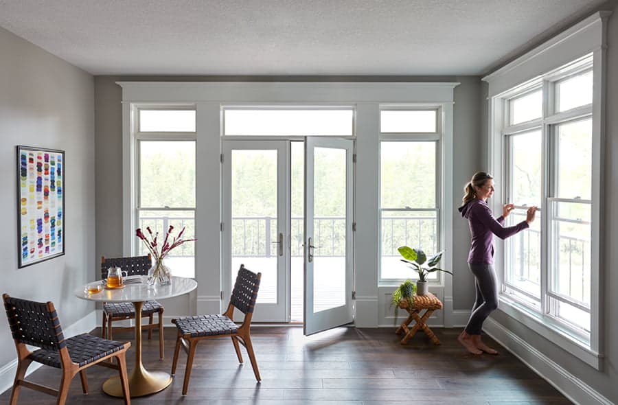 A woman in purple is opening one of two white double-hung windows that are near an open patio door