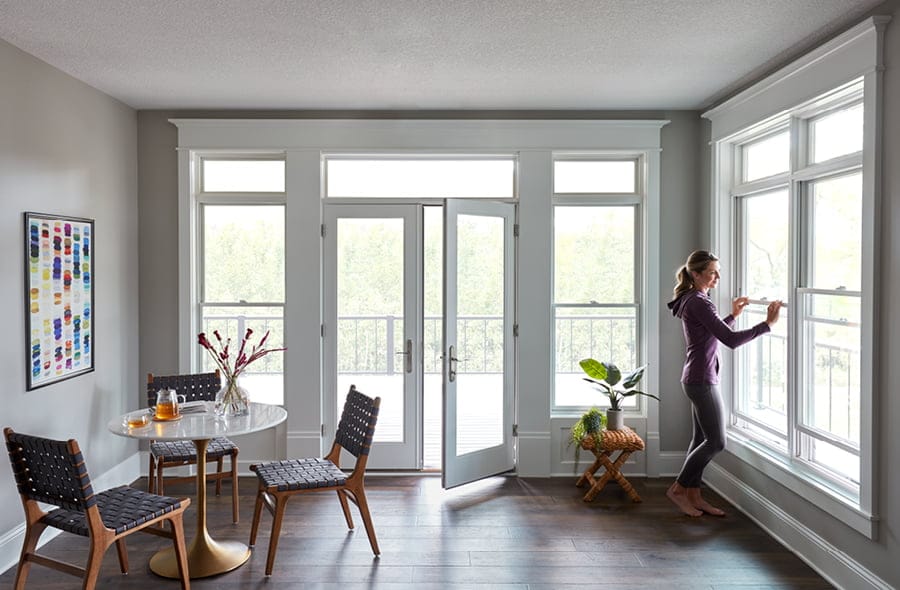 A woman in purple is opening one of two white double-hung windows that are near an open patio door