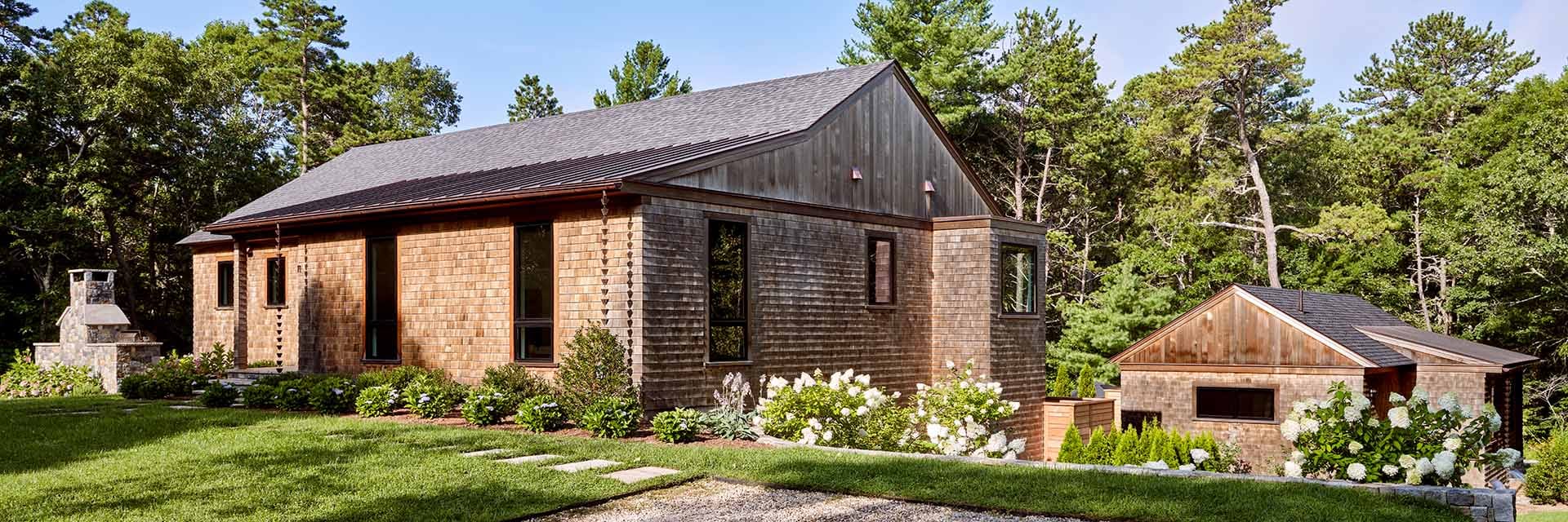 Cedar-shingle modern house with landscaped lawn, stone outdoor fireplace, hydrangea beds and detached garage surrounded by trees