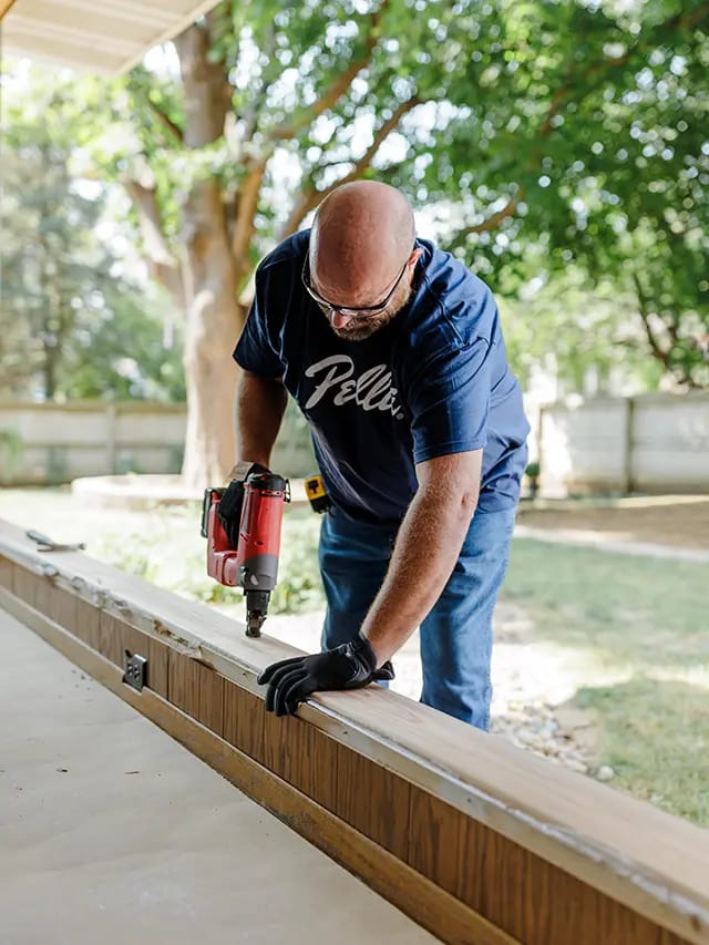Man using a red nail gun to install wooden porch trim during a backyard home improvement project.