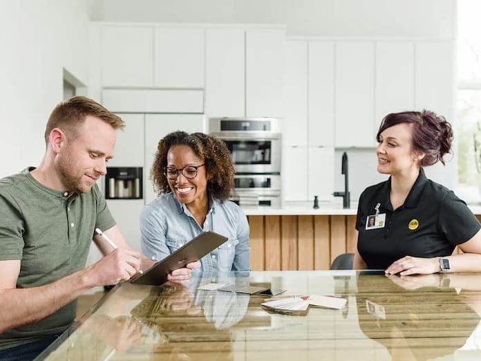 In-home consultation in a modern white kitchen with homeowners and a service professional reviewing a clipboard and documents.