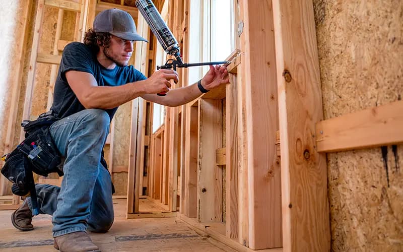 Kneeling construction worker uses a spray foam gun to seal a wooden window frame in new home wood stud framing