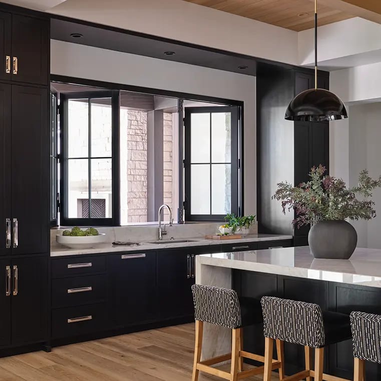 Modern kitchen with dark cabinets and marble countertops, island with black pendant light, three patterned bar stools, open black-framed windows and wood floor