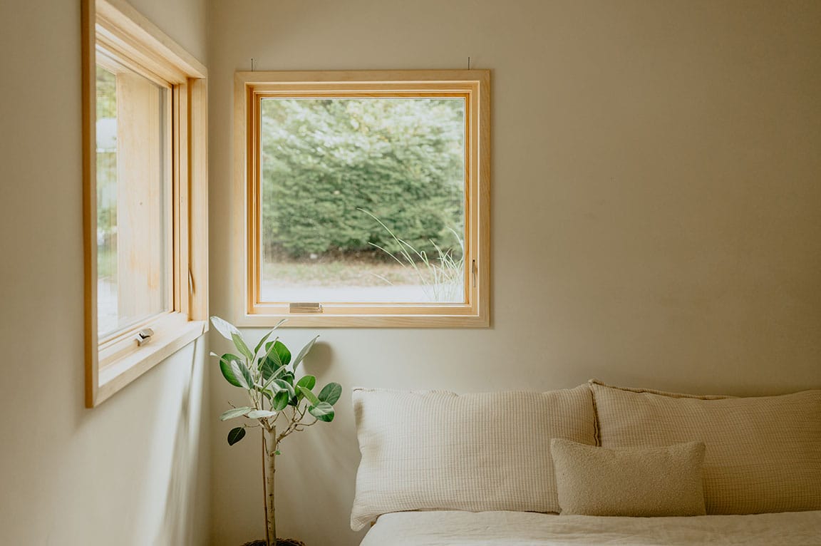 Bedroom interior with a wood‑framed window, soft neutral decor, and natural light overlooking greenery.