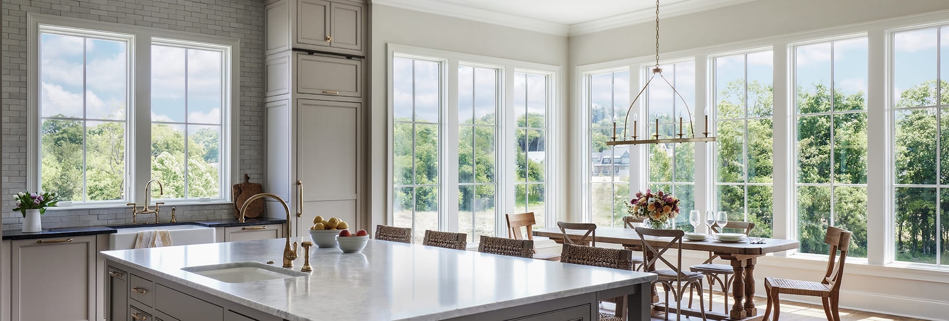 Bright farmhouse kitchen and dining area with marble island, brass faucets, wooden table, woven chairs and large pane windows overlooking lush trees.