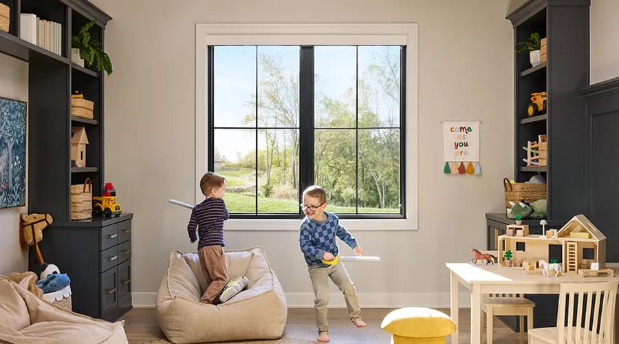 Two children perform playful activities with toys in a bright room featuring a large window and organized shelves.