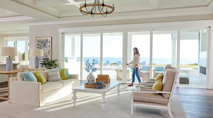 Bright coastal living room with sliding glass doors opening to ocean view, white sofa, coffee table, armchairs, and a woman walking toward the patio.