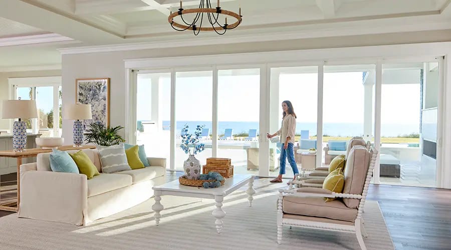 Bright coastal living room with sliding glass doors opening to ocean view, white sofa, coffee table, armchairs, and a woman walking toward the patio.