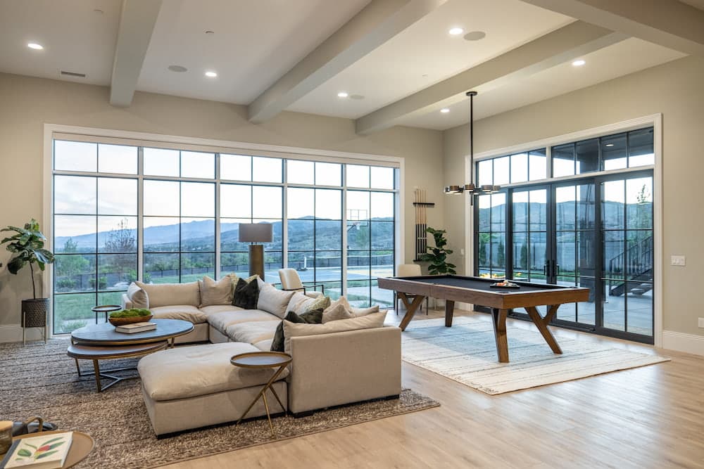 A walkout basement with a wall of picture windows behind a sectional and pool table and next to sliding glass doors.
