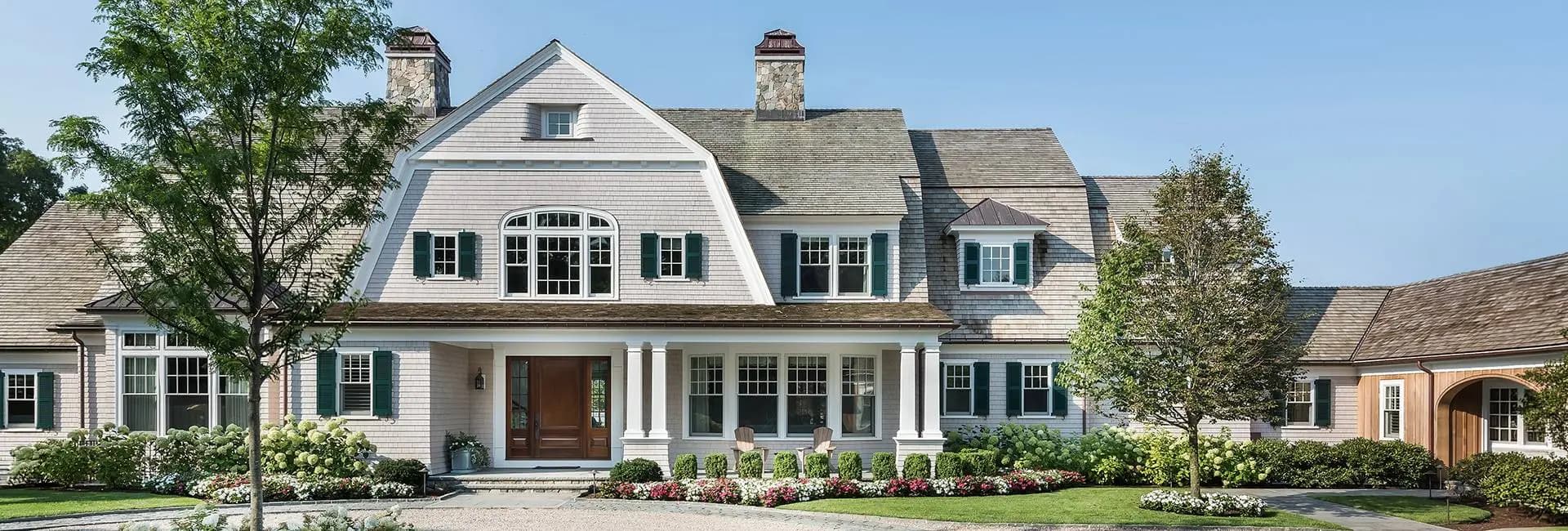 Shingle-style luxury home exterior with white siding, green shutters, central porch with columns, double chimneys and manicured flowerbeds