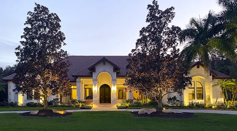 Mediterranean-style luxury home at dusk with arched entry, landscaped front yard, palm trees, and warm exterior lighting.