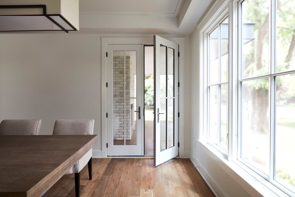 Wood French Doors With Custom Grille Pattern White farmhouse style dining room with wood flooring, double-hung windows on the right, and a set of white french doors open