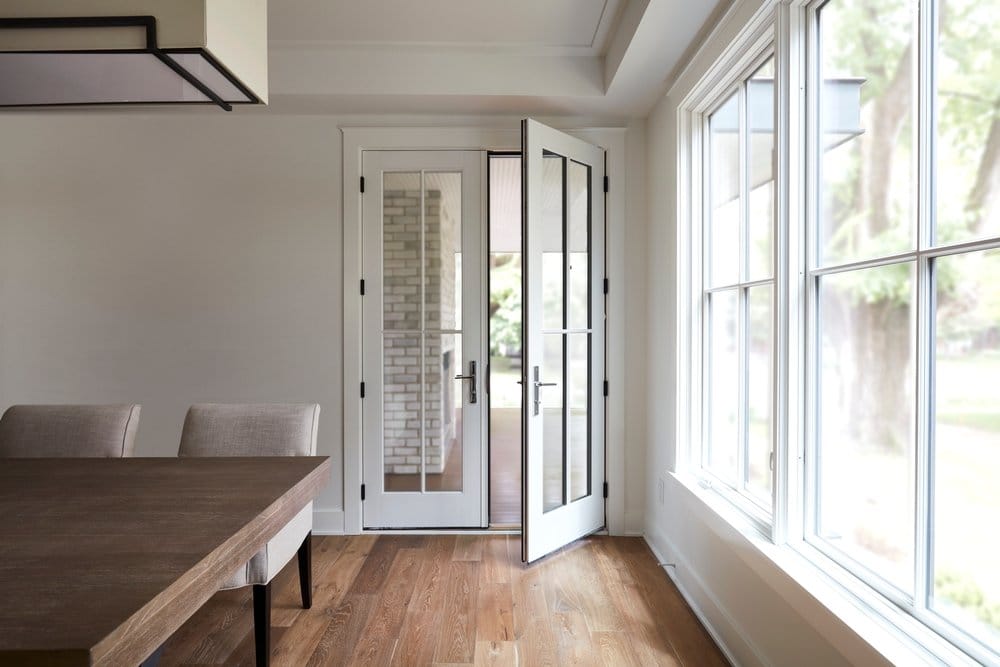 Wood French Doors With Custom Grille Pattern White farmhouse style dining room with wood flooring, double-hung windows on the right, and a set of white french doors open