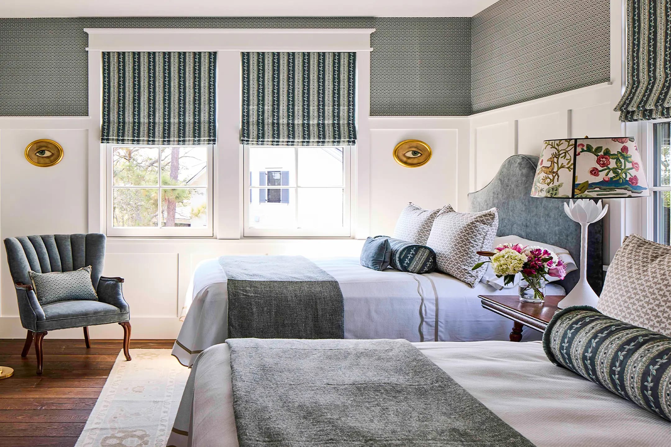 Guest bedroom with two twin beds, gray-blue linens, upholstered headboard, striped Roman shades, velvet armchair and floral table lamp