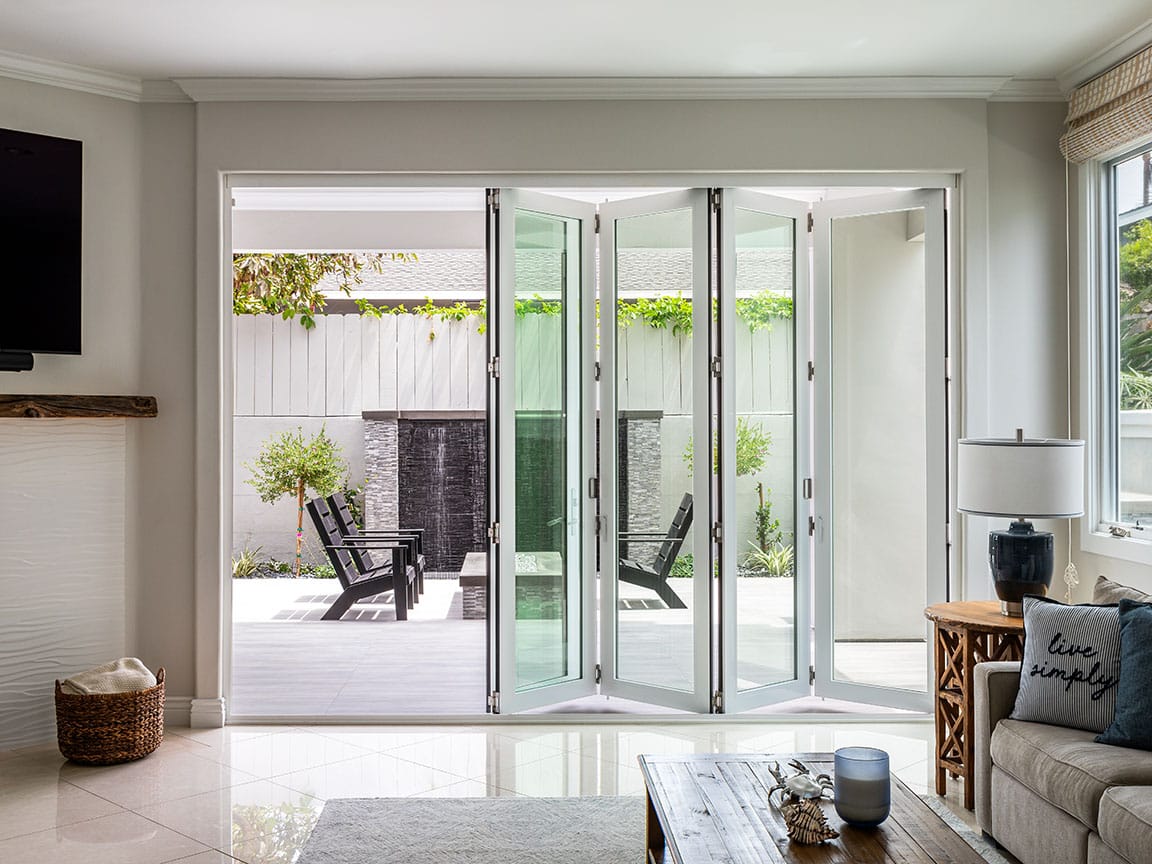 Large white bifold patio doors separate the minimalist living room from the exterior patio with greenery along the fence.