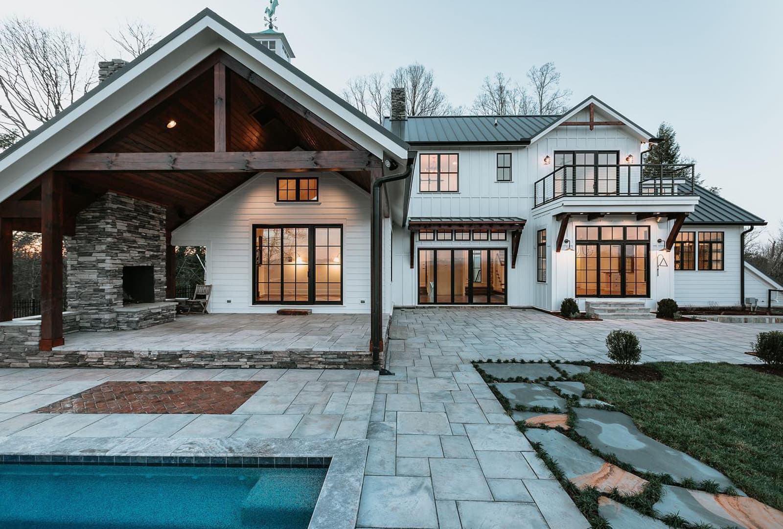 The exterior of a white farmhouse-style home with black windows and patio doors that overlook outdoor fireplace and pool.