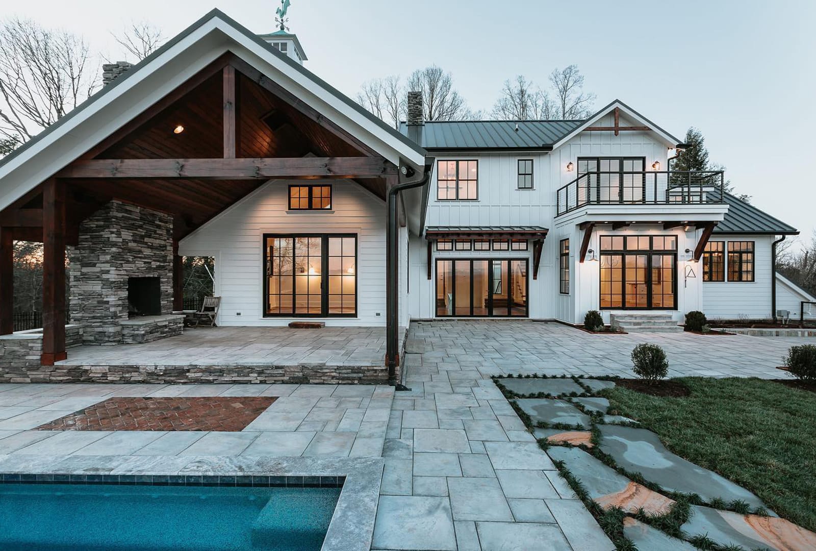The exterior of a white farmhouse-style home with black windows and patio doors that overlook outdoor fireplace and pool.