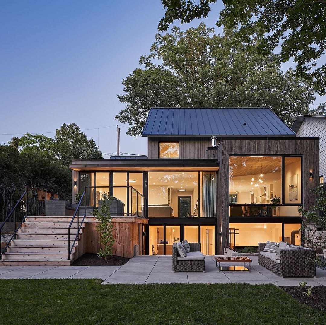 The exterior of a glass house features expansive basement windows and patio doors that connect to an outdoor living room.