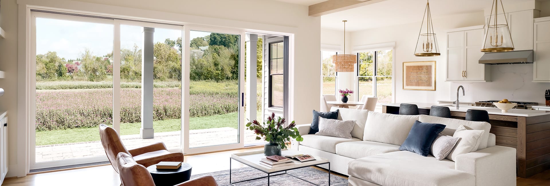 Bright open-concept living room and kitchen with large sliding glass doors, neutral sectional, leather chairs, island with pendant lights, and grassy field view
