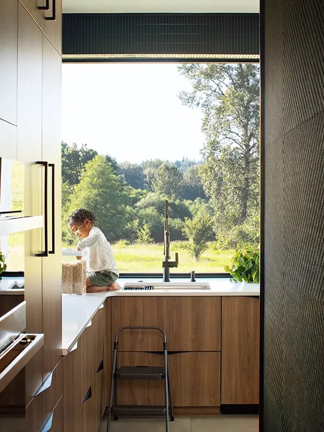 Child seated on white countertop by large picture window in modern kitchen with wood cabinets, black faucet, step stool and countryside view