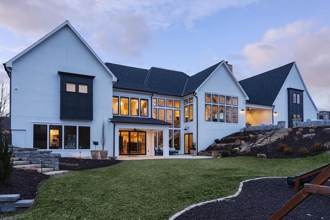 The backside of a white home shows a staircase with floor-to-ceiling windows and basement doors leading to a patio.