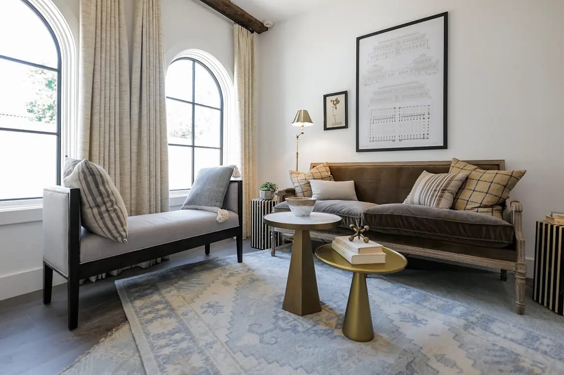 Modern cozy living room with arched windows and neutral curtains, gray chaise lounge, velvet sofa with patterned pillows, brass nesting side tables on a blue patterned rug.