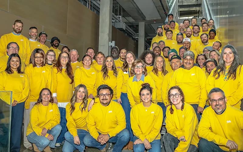 Group photo of employees wearing matching yellow sweatshirts posed on an indoor office stairway and lobby.